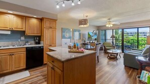 A modern kitchen with wooden cabinets, a dishwasher, and a dining area with a table and chairs. - Partial ocean view Kihei 1 bedroom condo, AC, walk to beach, shops, & dining (Kihei)
