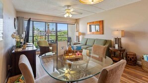 A modern living room with a glass table, a ceiling fan, and a view of greenery outside. - Partial ocean view Kihei 1 bedroom condo, AC, walk to beach, shops, & dining (Kihei)