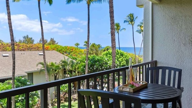 A balcony with a table and chairs, overlooking a tropical landscape with palm trees and a view of the ocean. - Partial ocean view Kihei 1 bedroom condo, AC, walk to beach, shops, & dining (Kihei)