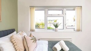 Ein Schlafzimmer mit Bett, Kissen, einem aufgerollten Handtuch, einem Fenster mit Vorhängen und einer Topfpflanze.