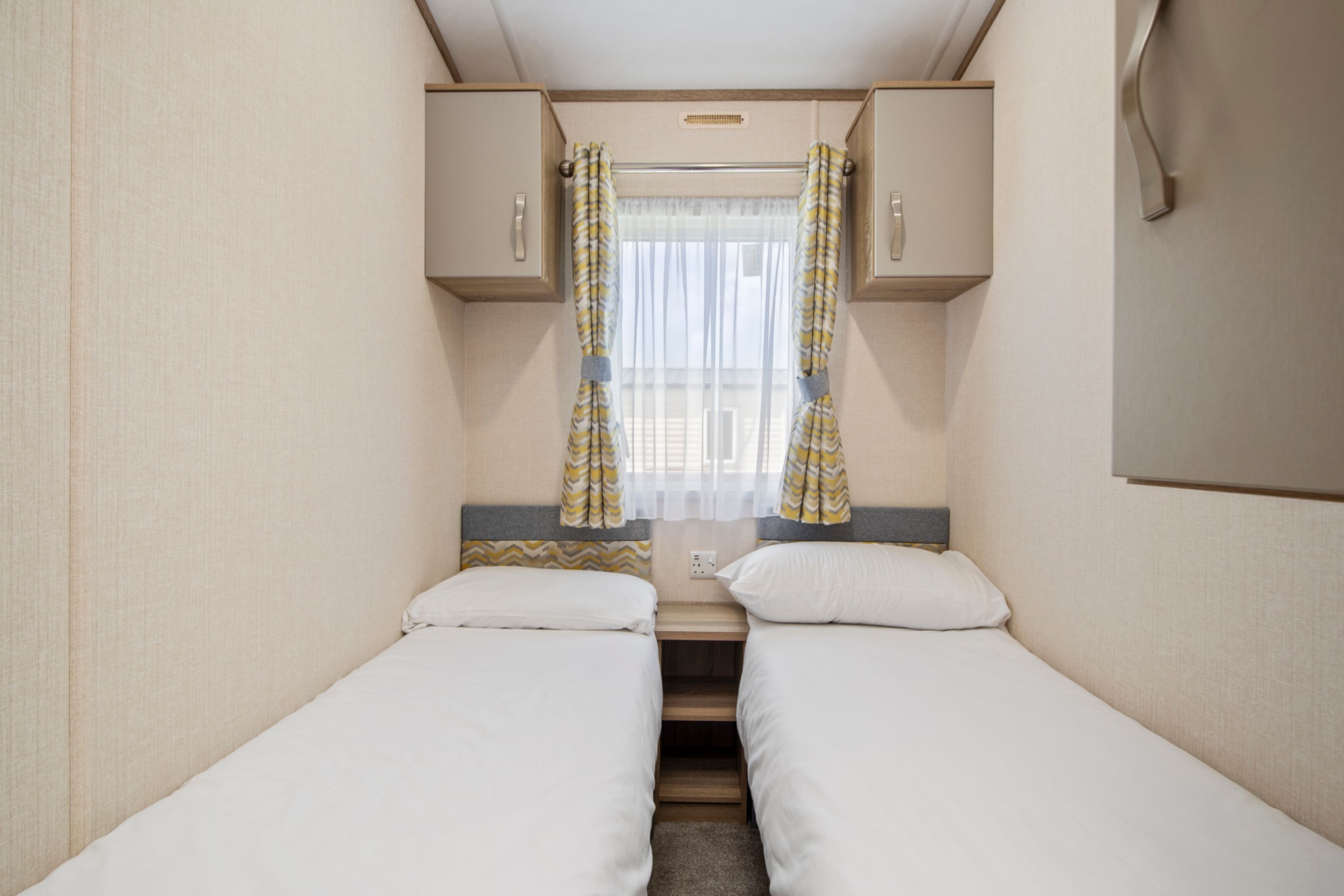 A compact bedroom with two single beds, a small window with curtains, and a built-in cabinet above the beds.