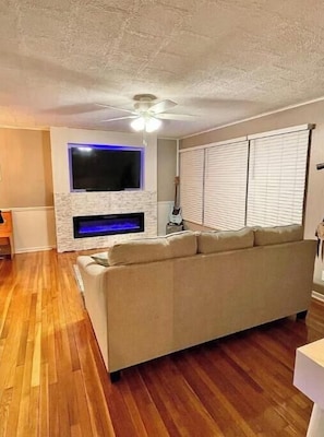 A living room with a beige sofa, a flat-screen TV mounted on the wall, a fireplace, and a ceiling fan.