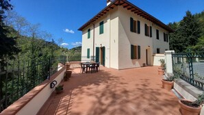 A two-story house with a tiled patio, green shutters, and a small table and chairs.