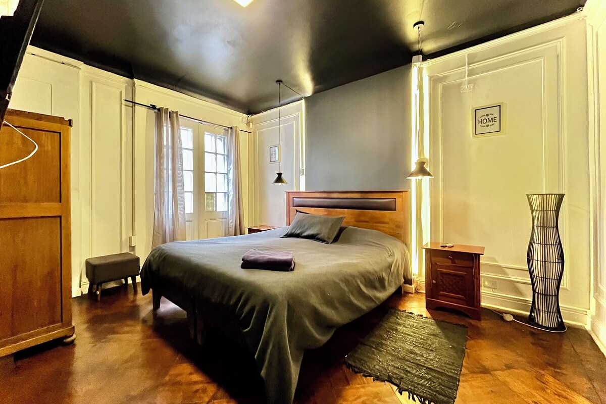Un dormitorio con una cama grande, una mesita de noche, una lámpara, un jarrón y una puerta.