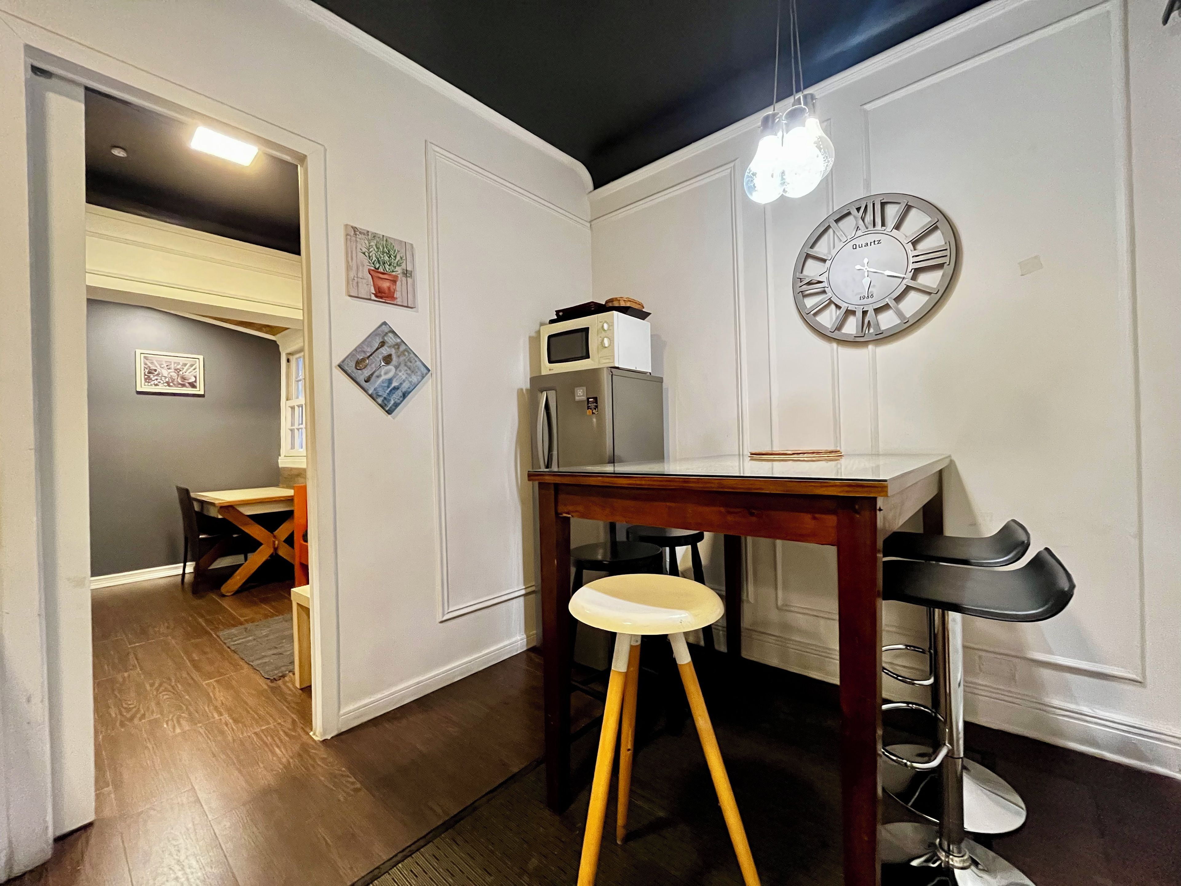 Una cocina compacta con una mesa de madera, un taburete, un refrigerador y un reloj de pared.