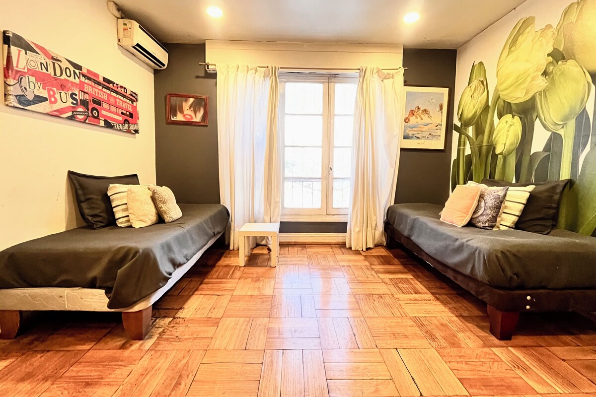 Una habitación con dos camas individuales, una mesita, un ventanal con cortinas y una pared con un cartel de viajes.