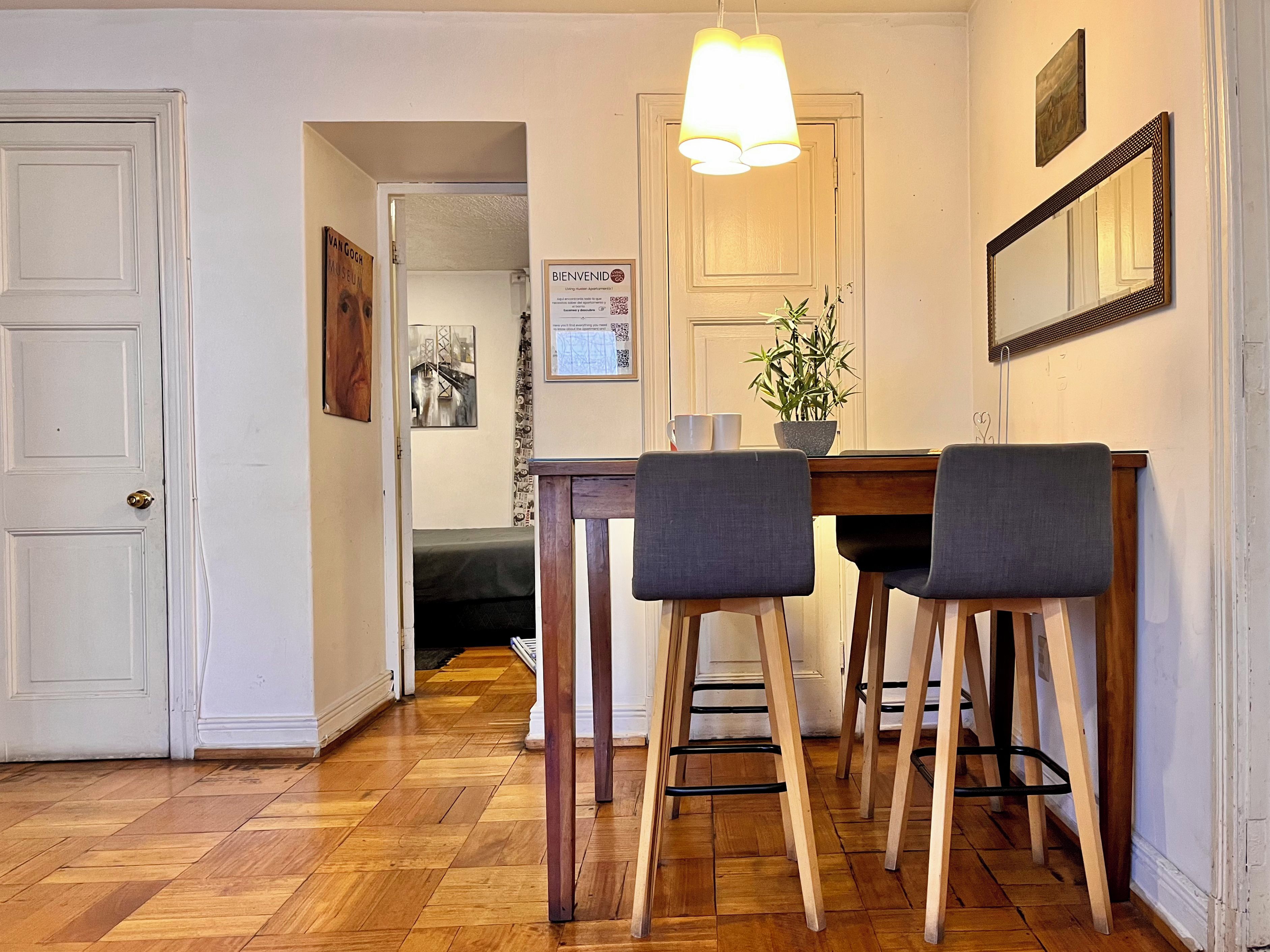 Un comedor con mesa de madera, dos sillas, una planta en maceta y una lámpara colgante.