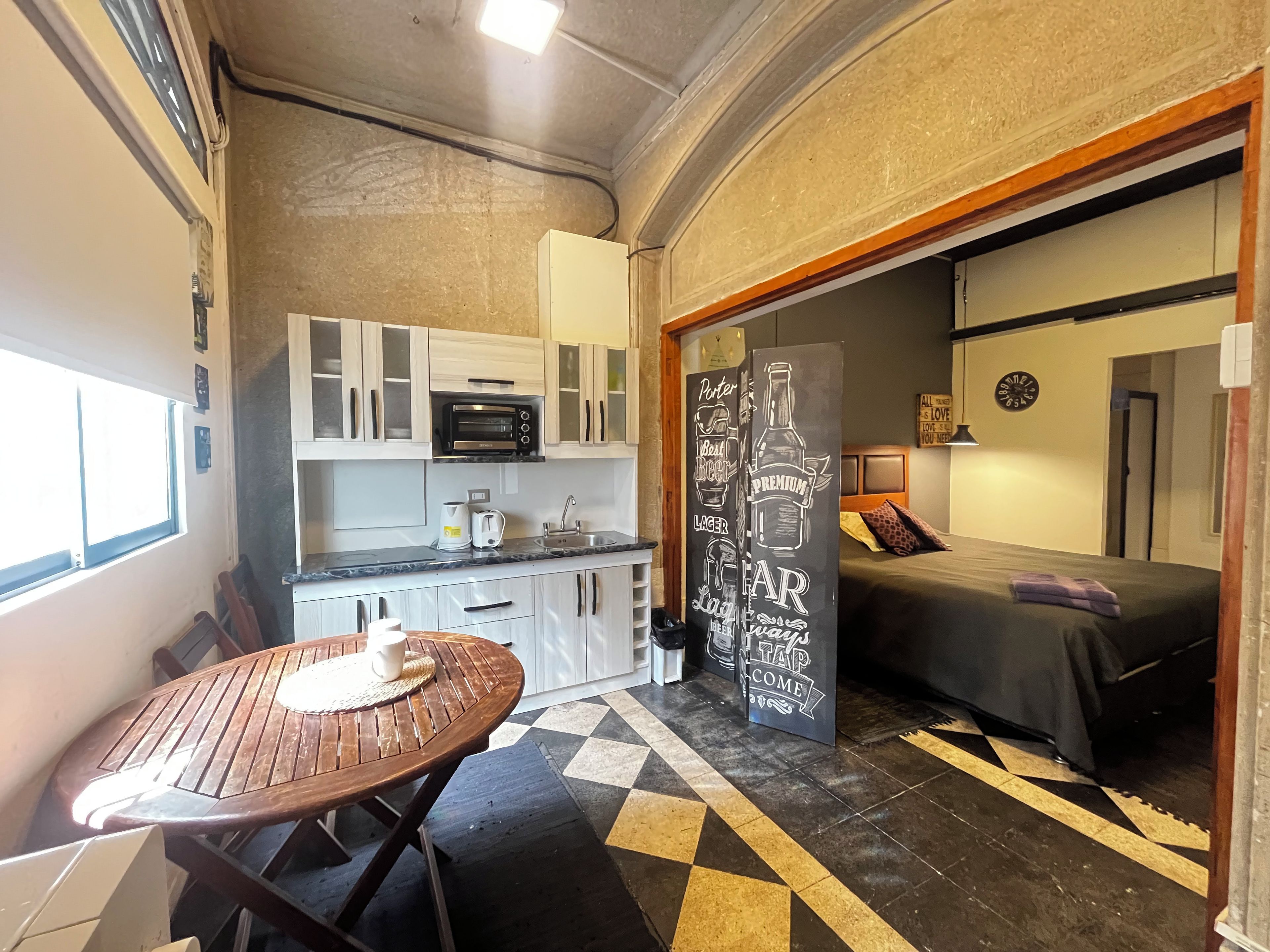 A compact kitchen with a microwave, toaster, and sink. There is a small dining table with a placemat. The room has a checkered floor pattern and a bed with a dark bedspread.