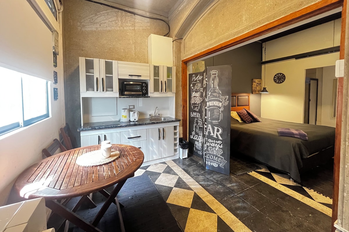 Cocina compacta con microondas, tostadora y lavabo. Hay una pequeña mesa de comedor con un mantel individual. El cuarto tiene un piso con diseño de cuadros y una cama con una colcha oscura.