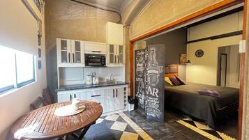 A compact kitchen with a microwave, toaster, and sink. There is a small dining table with a placemat. The room has a checkered floor pattern and a bed with a dark bedspread.