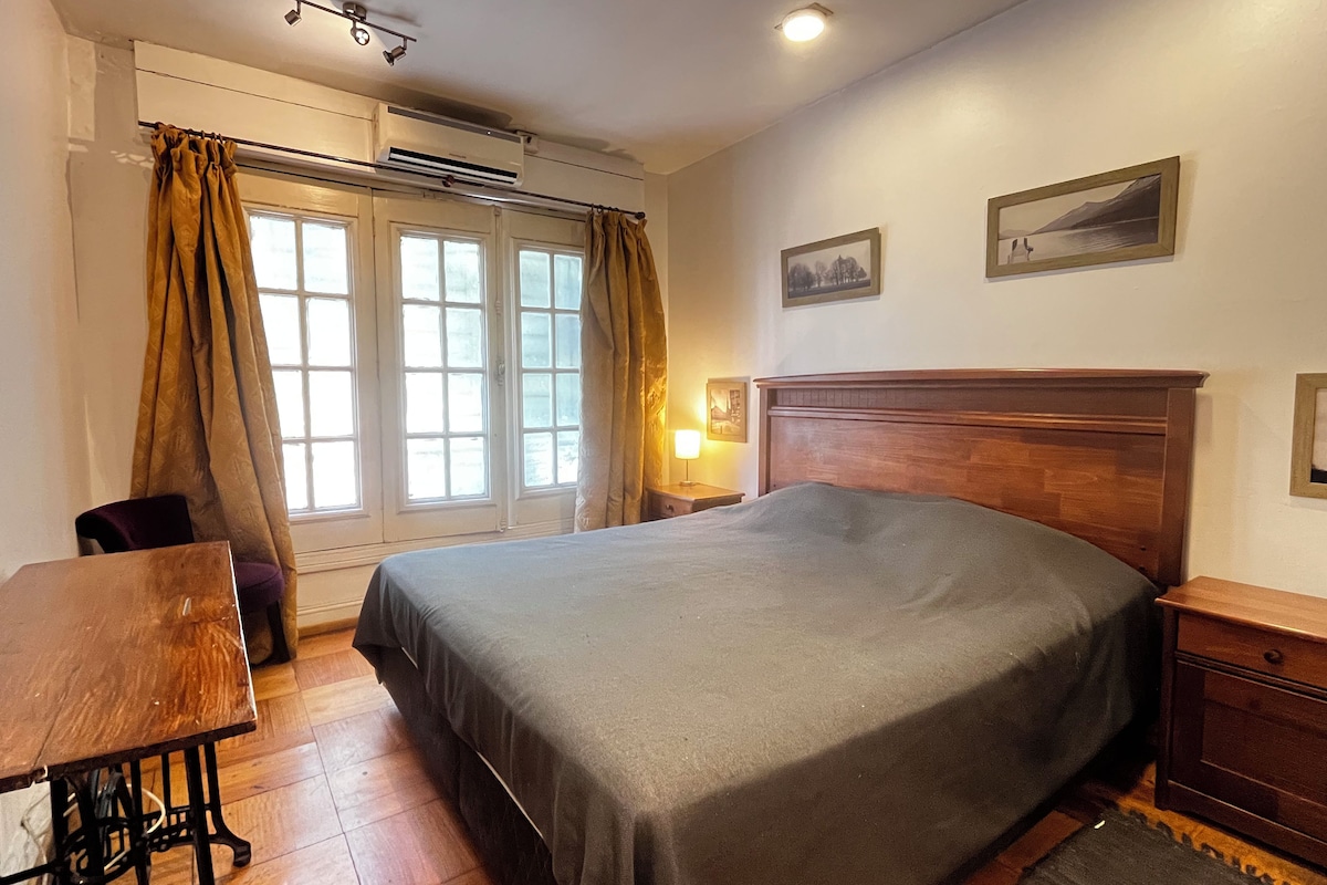 Habitación de hotel con cama, una mesa, una silla, aire acondicionado y ventana con cortinas.
