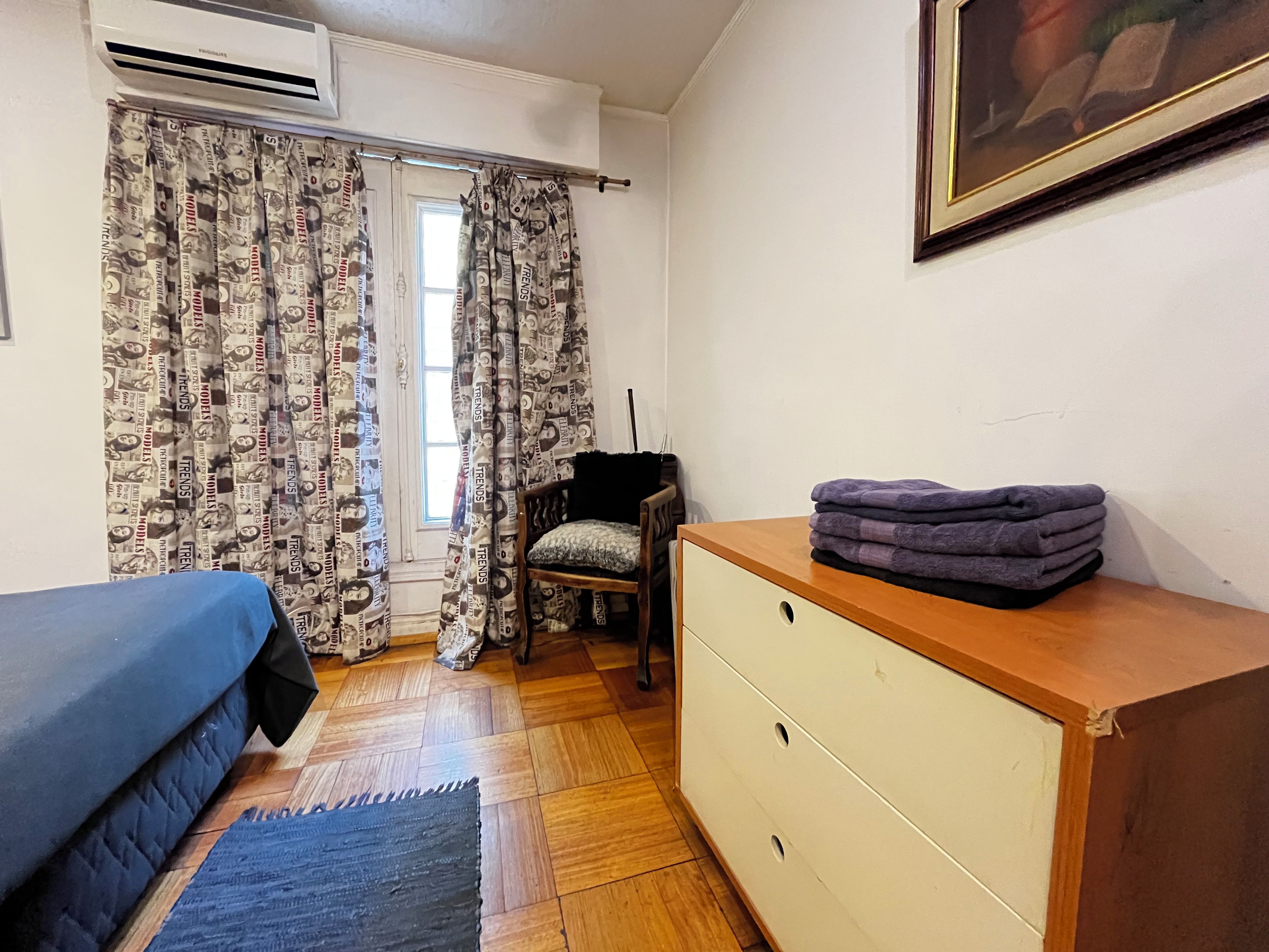 Una habitación con cama, cómoda, silla y ventana con cortinas estampadas.