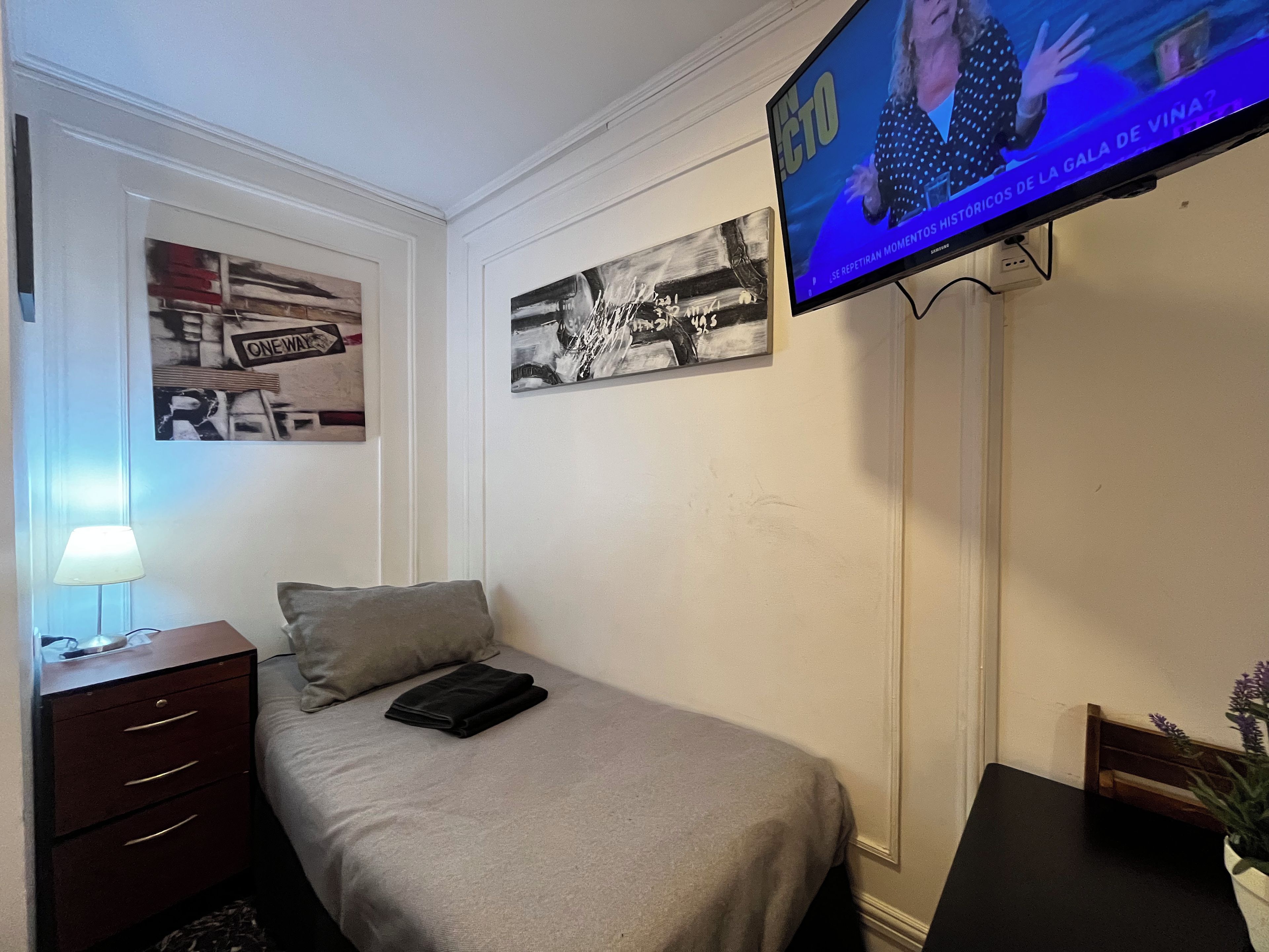 Una habitación de hotel con una cama, un televisor montado en la pared, una mesita de noche con una lámpara y dos cuadros enmarcados.