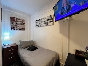 Una habitación de hotel con una cama, un televisor montado en la pared, una mesita de noche con una lámpara y dos cuadros enmarcados.