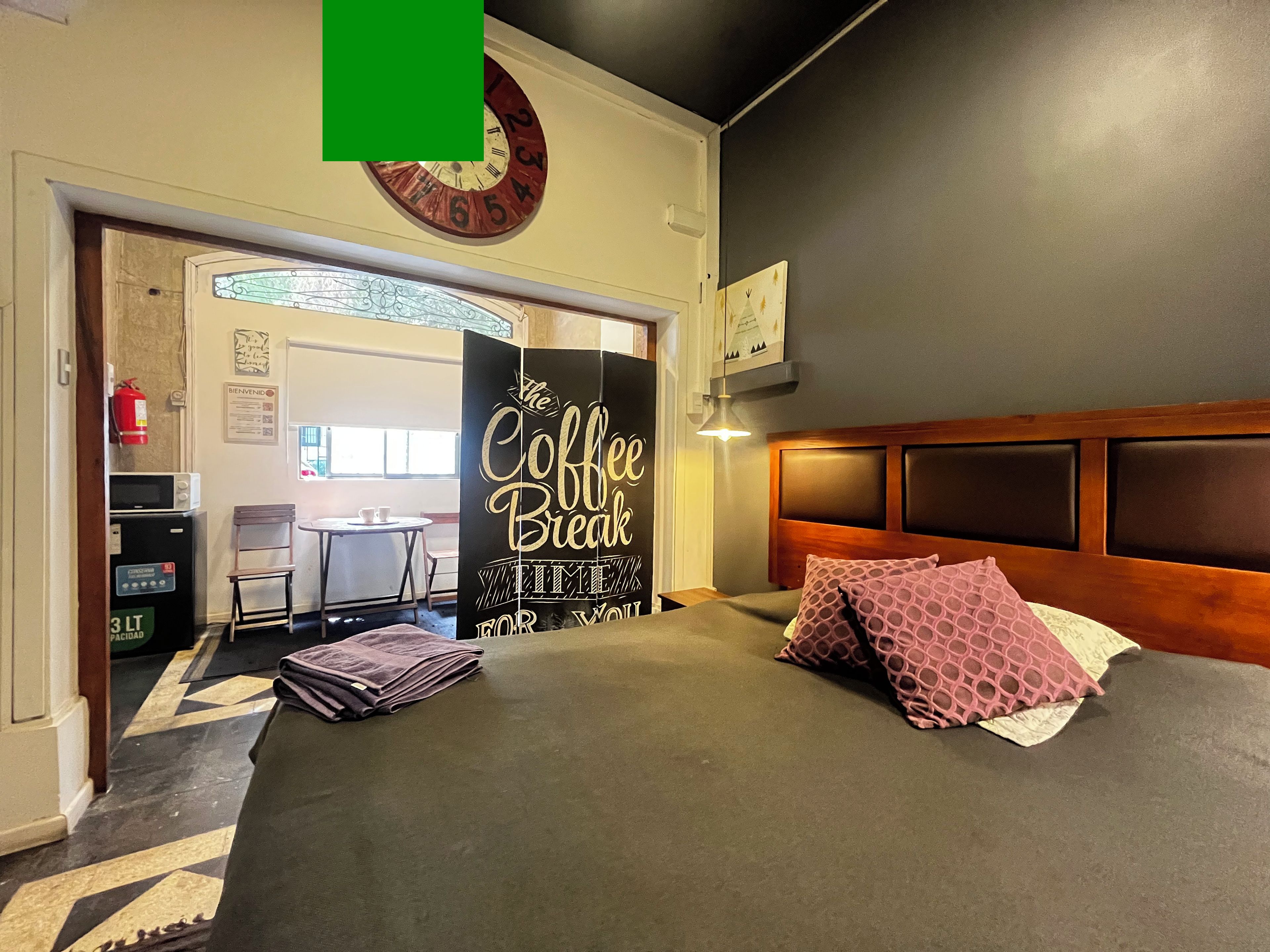 Una habitación de hotel con una cama, un cuadro con motivos de café, un reloj y una pequeña mesa con sillas.