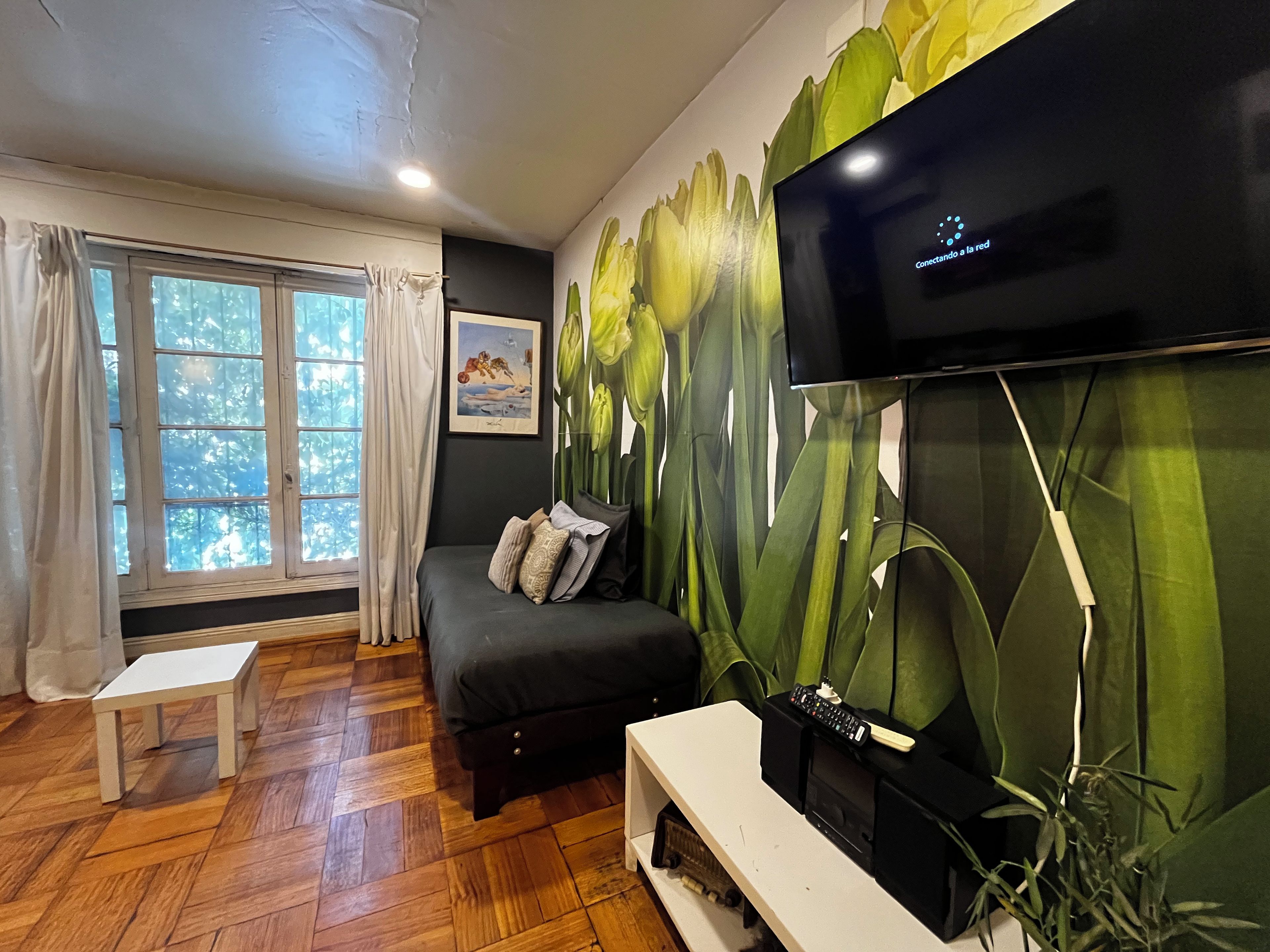 A room with a bed, a television mounted on the wall, a window with curtains, and a painting on the wall.