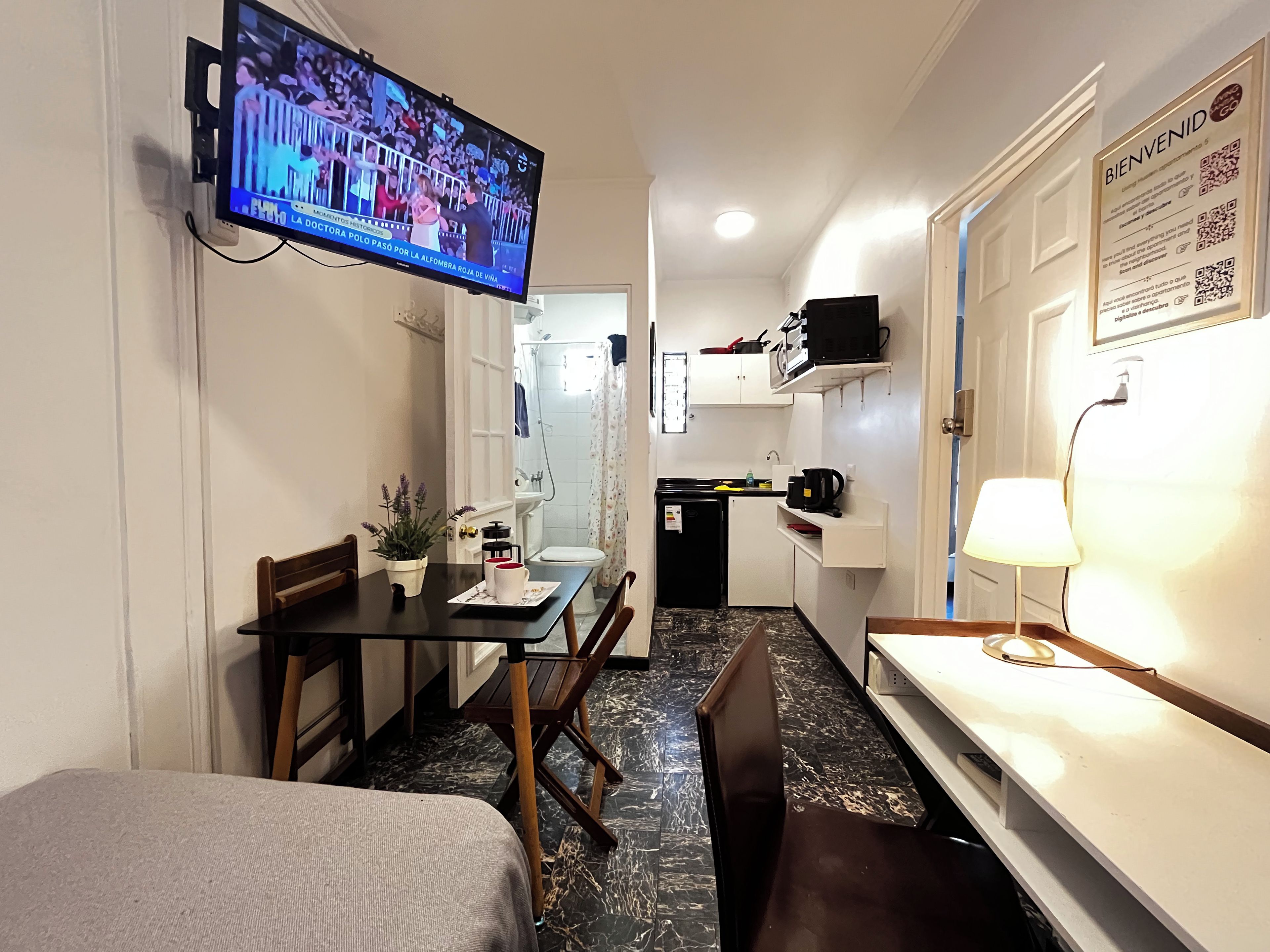 A compact hotel room with a television mounted on the wall, a small dining table with chairs, a bed, a kitchenette with a refrigerator, and a bathroom visible through an open door.