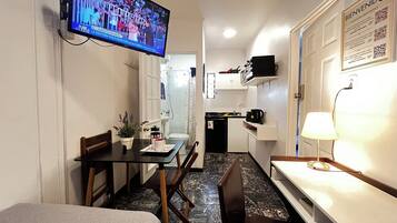 A compact hotel room with a television mounted on the wall, a small dining table with chairs, a bed, a kitchenette with a refrigerator, and a bathroom visible through an open door.