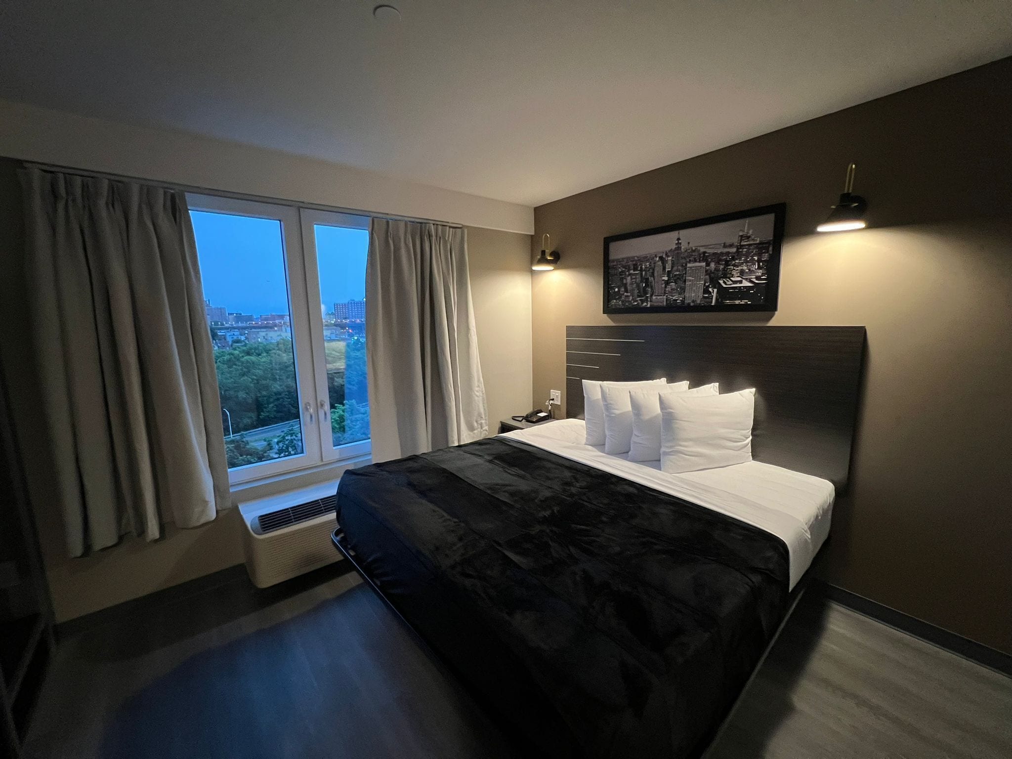 A modern hotel room with a large bed, a city view, and a framed picture on the wall.