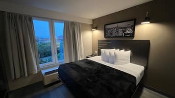 A modern hotel room with a large bed, a city view, and a framed picture on the wall.