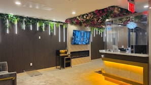A modern reception area with a wooden panel wall, a TV mounted on the wall, a reception desk with a glass top, and a decorative floral arrangement above the desk. - LIV Hotel Bronx - Newly Built (Bronx)