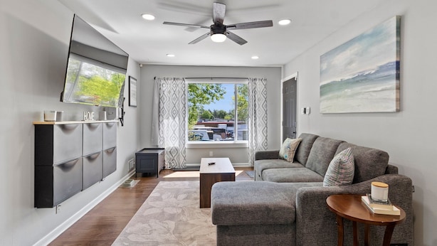 A modern living room with a grey sectional sofa, a wooden coffee table, a TV mounted on the wall, and a ceiling fan. - MKE 2 Bed W/ PKNG 15M Downtown (Milwaukee)