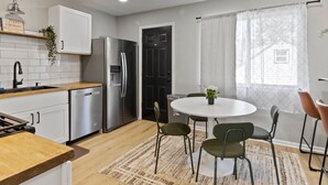 A modern kitchen with white cabinets, stainless steel appliances, and a wooden countertop. There is a dining area with a round table and green chairs. - MKE 2 Bed W/ PKNG 15M Downtown (Milwaukee)