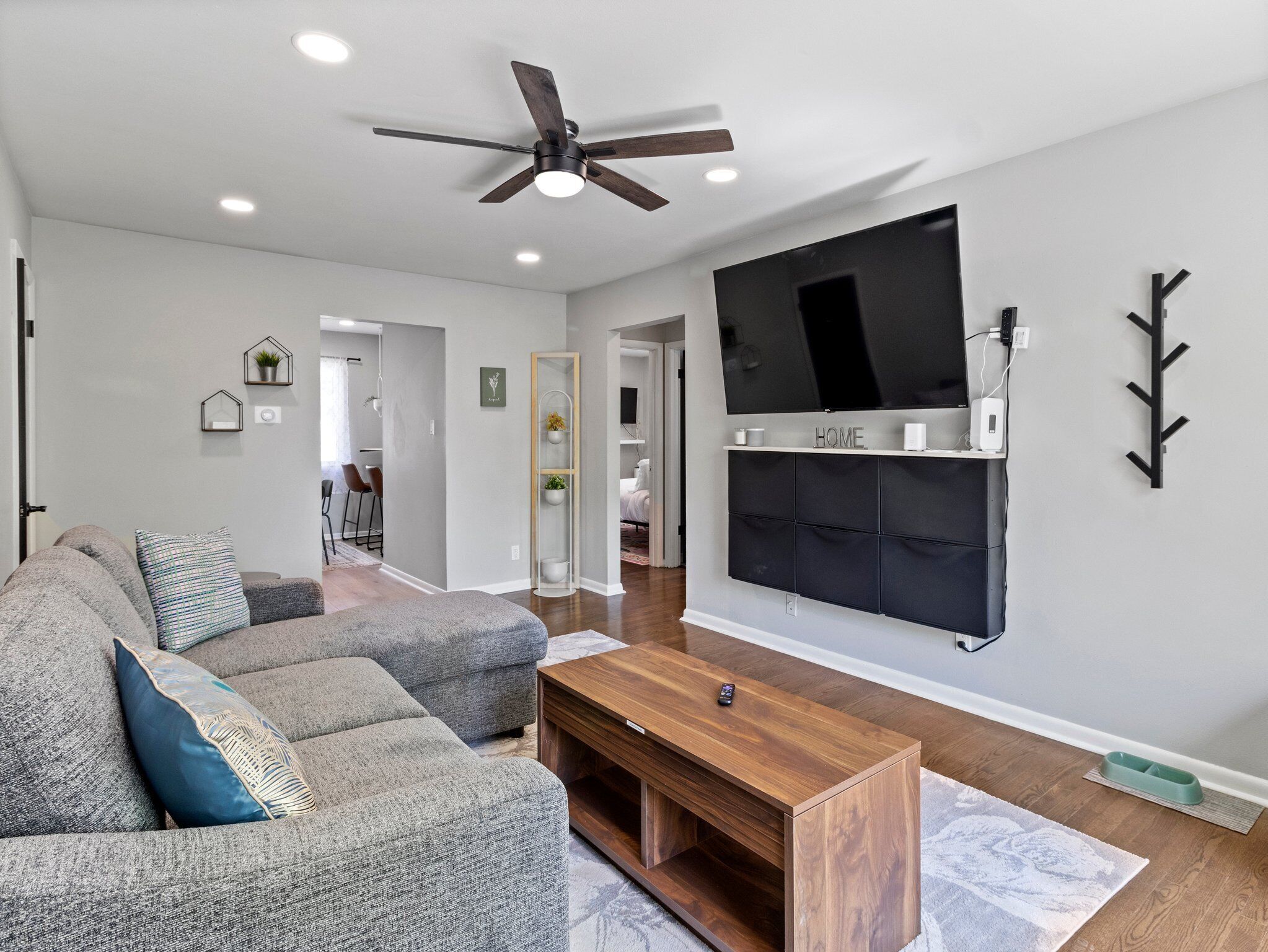 A modern living room with a sectional sofa, a wooden coffee table, a flat-screen TV mounted on the wall, and a ceiling fan.