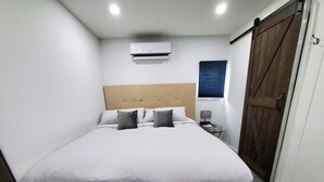 A neatly made bed with a headboard, a bedside table with a lamp, and an air conditioning unit above it. - Perfect Guest house/studio just 15mins drive to the beach for a relaxing getaway (West Park)