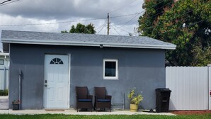 A small, single-story building with a gray exterior, a white door, and a small window. There are two wicker chairs with blue cushions outside, a potted plant, and a trash can. - Perfect Guest house/studio just 15mins drive to the beach for a relaxing getaway (West Park)