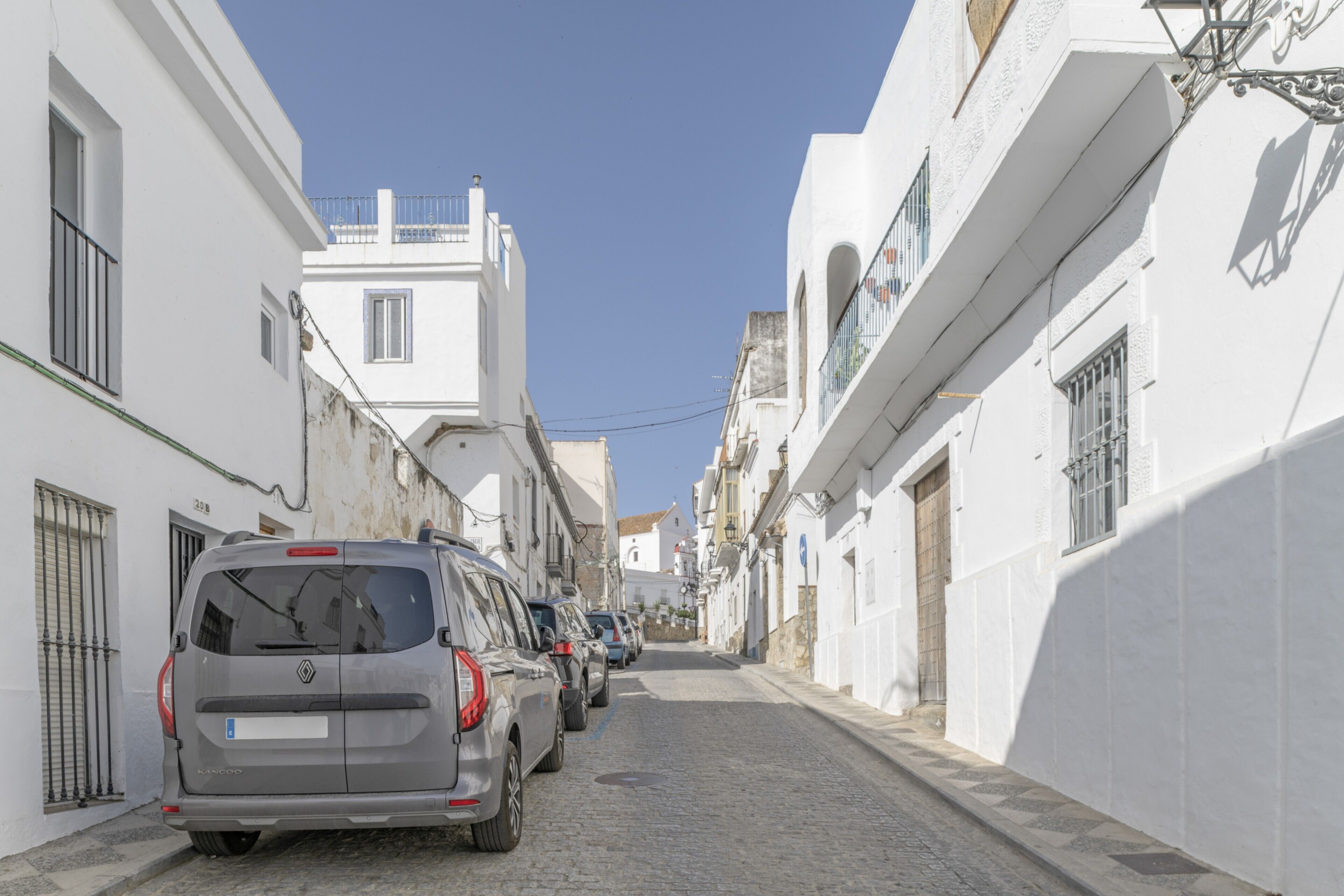 Une rue étroite bordée de bâtiments blancs, de voitures garées et d’un ciel bleu azur.