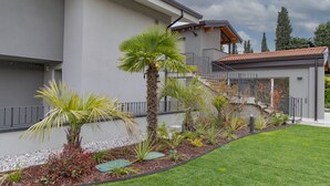 A modern building with a grey exterior, a well-maintained lawn, and a variety of plants including a palm tree. - Modern apartment with pool a few steps from Lake Garda (Padenghe Sul Garda)