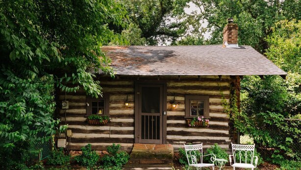 A log cabin with a wooden door, two windows, and a chimney. There are two white metal chairs and a small table in front of the cabin.