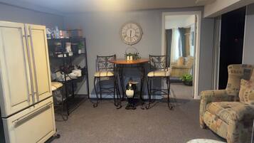 A compact living space with a kitchenette, a dining table with chairs, a decorative clock, and a patterned armchair.