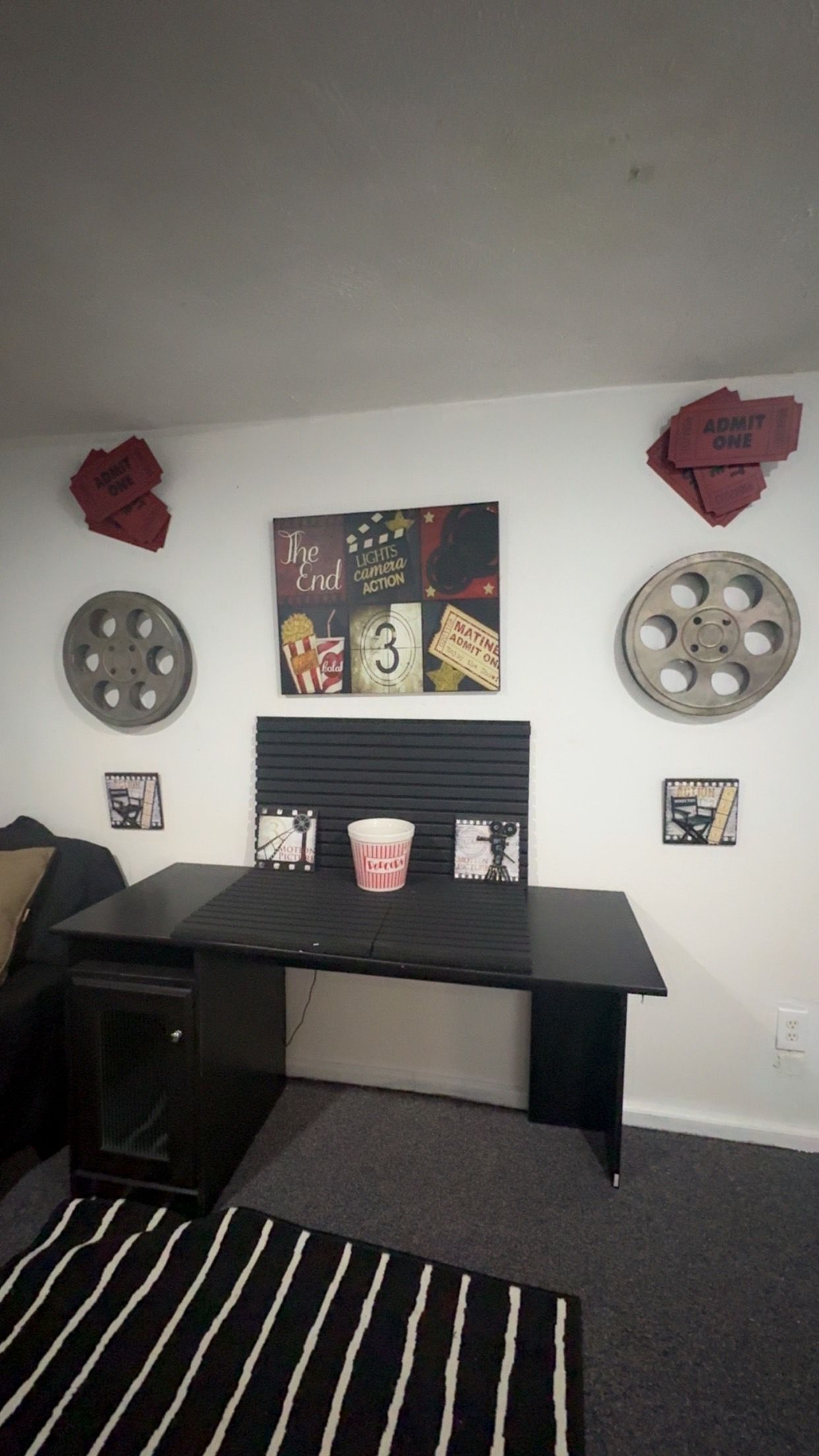 A room with a black desk, a striped rug, and a wall decorated with movie memorabilia including film reels and a poster.