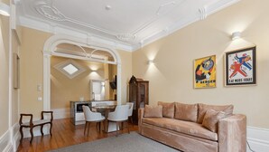 Una sala de estar con un sofá color beige, una mesa de comedor con sillas, una zona de cocina y una escalera que sube a un nivel superior.