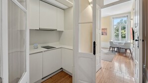 Et moderne køkken med lyse skabe, en indbygget komfur og en lille bordplade. En spiseplads med et bord og stole er synlig gennem en åben dør.