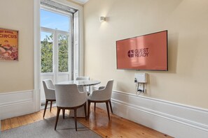 Une pièce avec une table ronde, quatre chaises, un téléviseur fixé au mur et une fenêtre donnant sur des arbres.