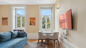 En stue med en blå sofa, et rundt bord med stole, to vinduer med plakater og et ophængt fjernsyn på væggen.