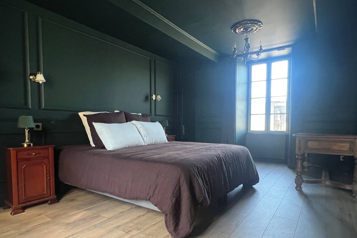 Une chambre avec un grand lit, un plancher en bois, une table de chevet avec une lampe, une commode et une fenêtre laissant entrer la lumière naturelle.
