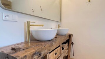 Un baño con dos lavamanos blancos tipo pedestal, un mueble de tocador de madera, un espejo grande y un grifo dorado.