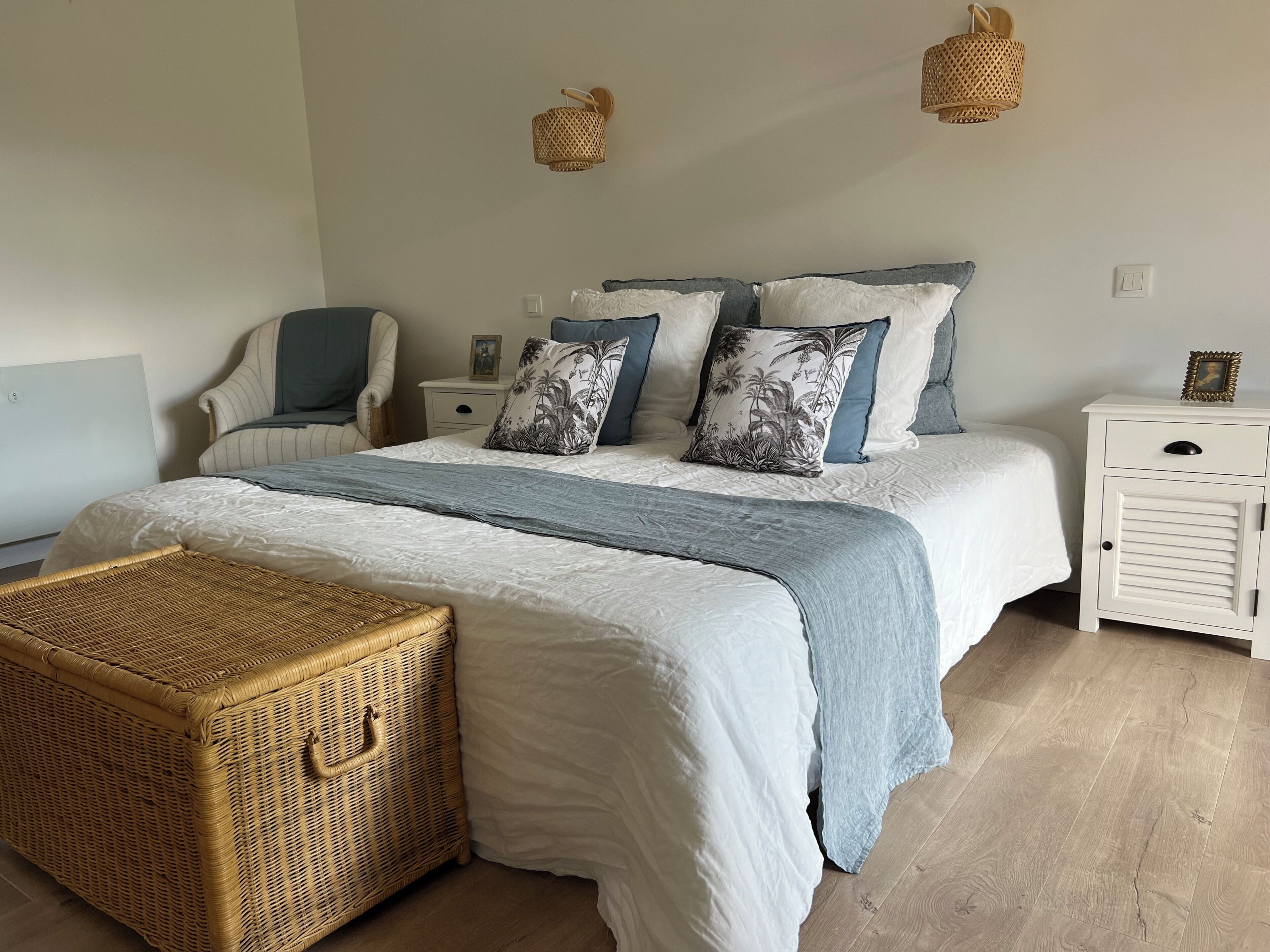 A neatly arranged bedroom with a bed, bedside tables, a chair, and a wicker chest.