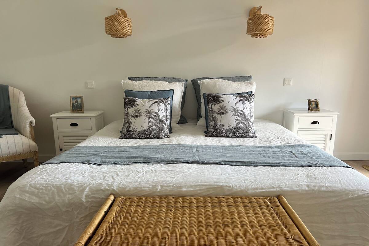 Un lit bien fait avec deux oreillers, une table de chevet ornée d’un cadre photo, une chaise en osier et deux lampes fixées au mur.