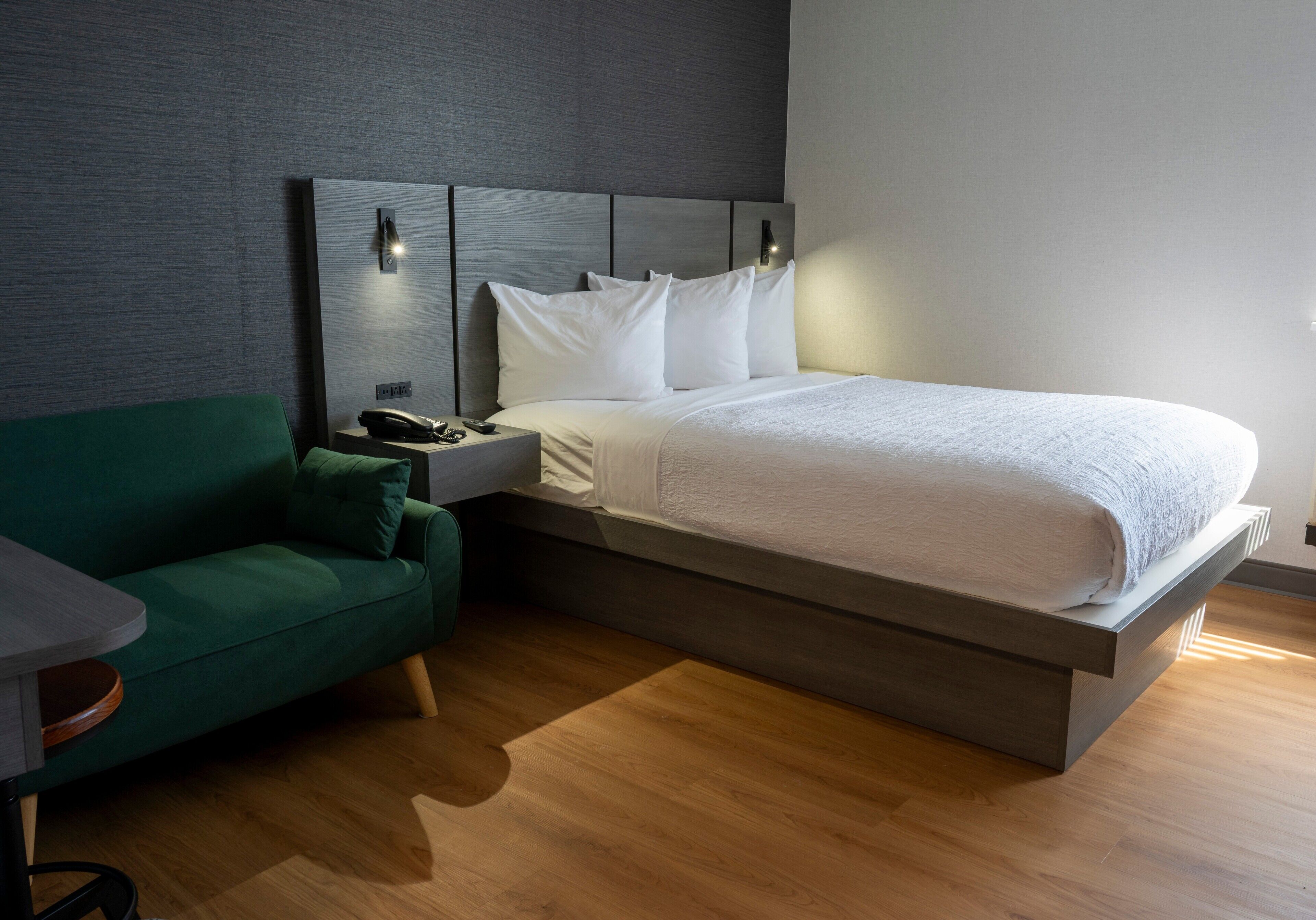 A modern hotel room with a large bed, a green sofa, a bedside table with a lamp, and a nightstand with a phone.