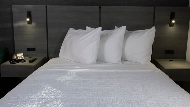 Ein modernes Hotelzimmer mit einem großen Bett, weißen Leinentextilien und einem Kopfteil mit integrierter Beleuchtung. An den Nachttischen befinden sich ein Telefon und eine Fernbedienung.