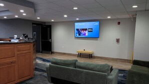 Eine Hottelobby mit einer Rezeption, einem großen Flachbildfernseher an der Wand, einem grünen Sofa und einer kleinen Bank.
