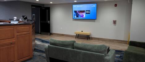A hotel lobby with a reception desk, a flat-screen TV mounted on the wall, a bench, and green upholstered seating.