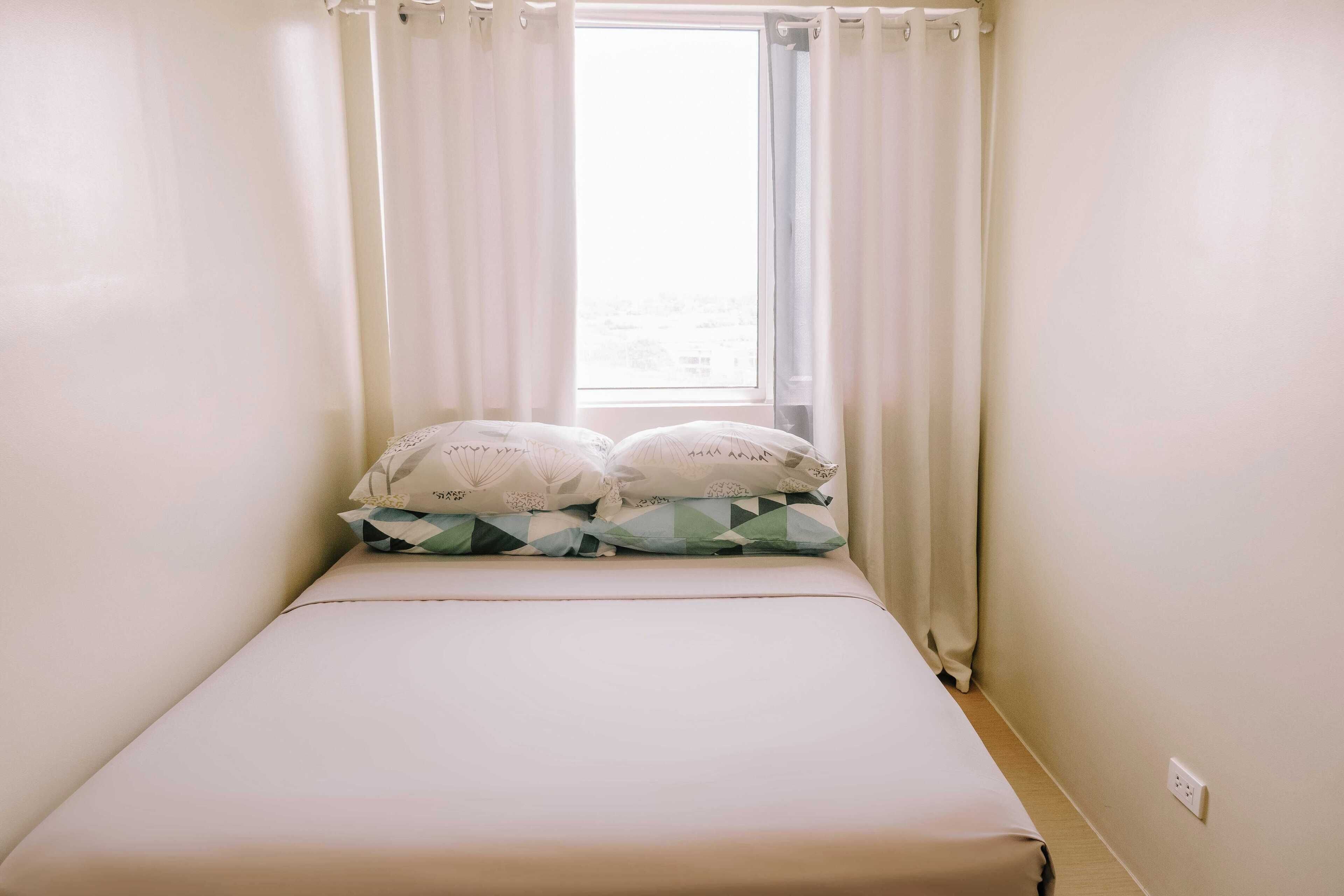 Un letto piccolo e ben rifatto con materasso bianco e due cuscini, una finestra con tende bianche e un interruttore a parete.