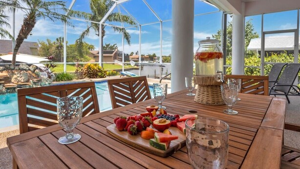 Une table en bois, sur laquelle sont disposés un plateau de fruits et des verres, donnant sur une piscine et des palmiers.