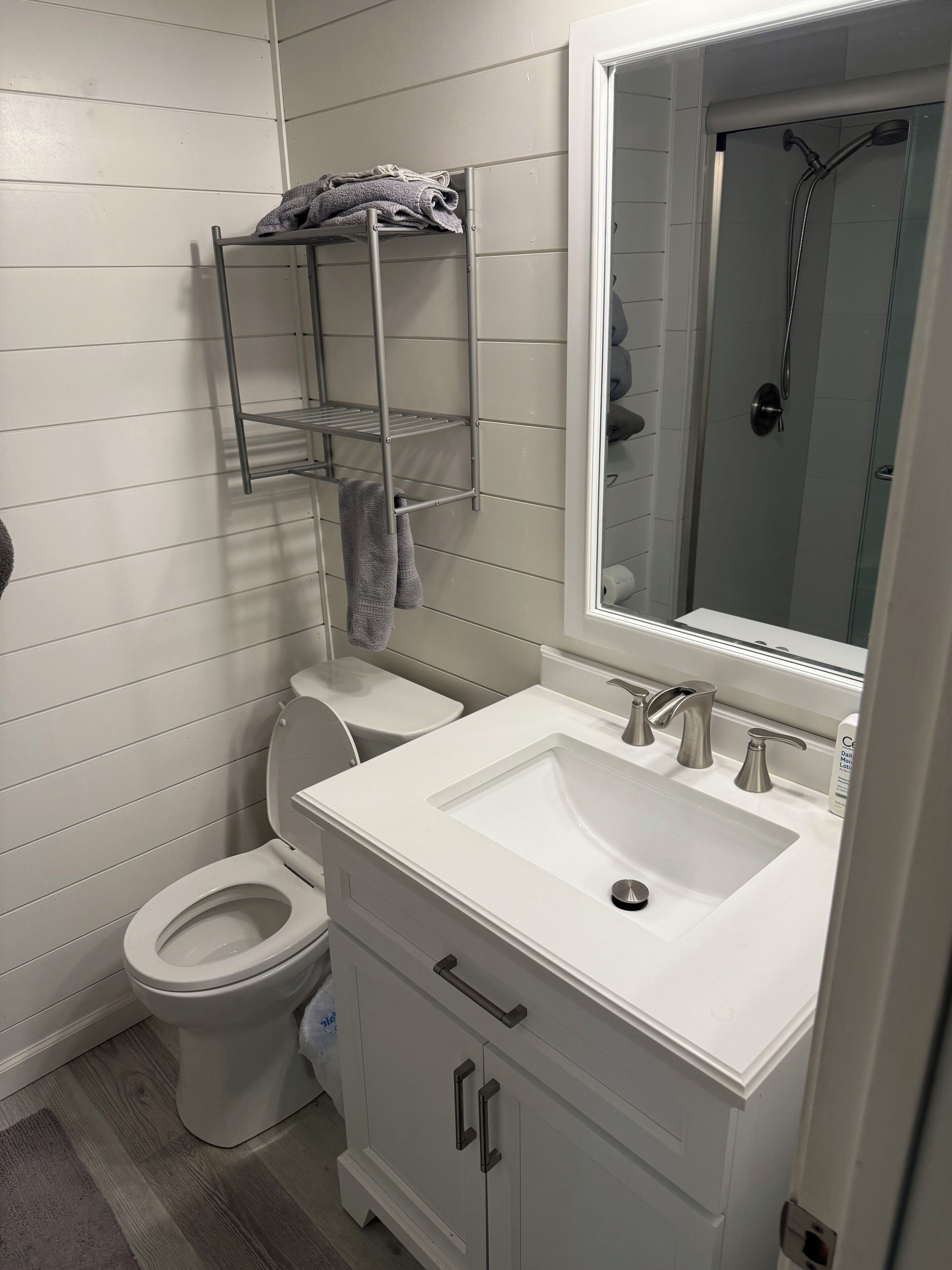 Ein Badezimmer mit einem weißen Waschbecken, einer Toilette und einer Duschkabine. Über der Toilette befindet sich ein Handtuchhalter und eine Ablage mit Handtüchern.